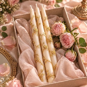 May include: Three off-white taper candles decorated with metallic gold rose and vine embellishments. The candles are arranged vertically inside a rectangular box lined with soft, light pink fabric, accompanied by several pink rose blossoms.