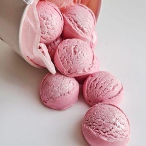 May include: A white paper cup tipped over, spilling out several scoops of pink ice cream. The ice cream has a textured surface and a slight shimmer. Pink tissue paper lines the cup.
