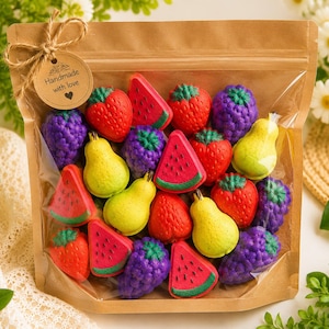 May include: A clear plastic pouch with a brown paper top contains assorted fruit-shaped bath bombs including red strawberries, purple blackberries, yellow pears, and watermelon slices. A round brown tag tied with twine reads Handmade with love.