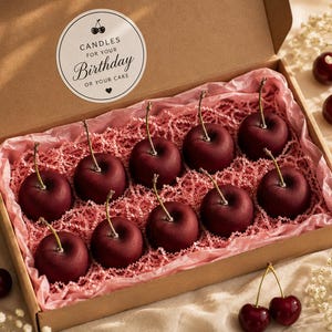 May include: A cardboard gift box containing ten dark red cherry-shaped candles nestled in pink crinkle paper. The inside lid features a circular white sticker with the text: CANDLES FOR YOUR Birthday OR YOUR CAKE. Two real cherries are visible nearby.