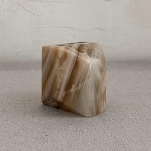 May include: A small, square, decorative candle holder. The candle holder is made of a stone material with a striped pattern of light brown and cream colors. The candle holder is sitting on a neutral-colored surface.