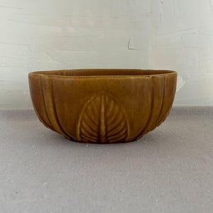May include: A brown ceramic bowl with a leaf design in the center. The bowl has a ribbed exterior and a wide, oval shape. The color is a warm, earthy tone, suitable for home decor.