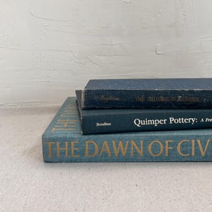 May include: Three vintage books stacked on top of each other. The top book is a dark blue, the middle book is a teal color, and the bottom book is teal with gold lettering that reads "THE DAWN OF CIV".