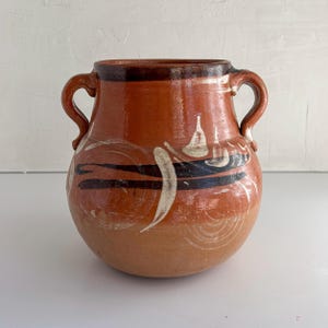 May include: A handcrafted, terracotta-colored ceramic vase with two curved handles. The vase features a dark brown rim and is adorned with white and black painted designs. The overall shape is round with a slightly wider base.