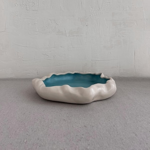 May include: A shallow ceramic bowl with a smooth, matte white exterior and a vibrant turquoise interior. The bowl features an irregular, wavy, and organic rim shape that creates a rippled effect around the entire circumference of the vessel.