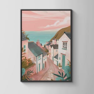 May include: A framed art print depicting a coastal village with pale pink and white houses lining a cobblestone path that leads towards the sea. The scene features a soft pink sky, dark grey roofs, teal doors, and lush green foliage in the foreground.