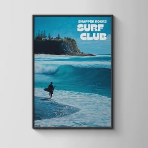 May include: A framed print featuring a surfer walking into the ocean with a surfboard. The artwork has a blue and white color scheme, with the text "SNAPPER ROCKS SURF CLUB" at the top. The image depicts a coastal scene with waves and a cliffside.