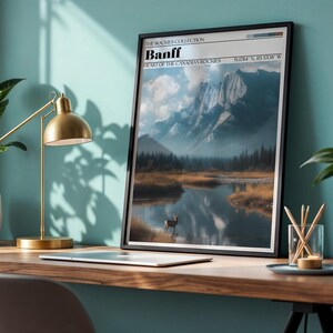 May include: A framed print featuring a scenic landscape of Banff National Park, with a mountain range, lake, and deer. The print has a black frame and includes the text "Banff, Heart of the Canadian Rockies". A gold desk lamp and a plant are also visible.