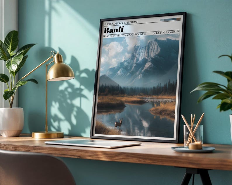 May include: A framed print featuring a scenic landscape of Banff National Park, with a mountain range, lake, and deer. The print has a black frame and includes the text "Banff, Heart of the Canadian Rockies". A gold desk lamp and a plant are also visible.