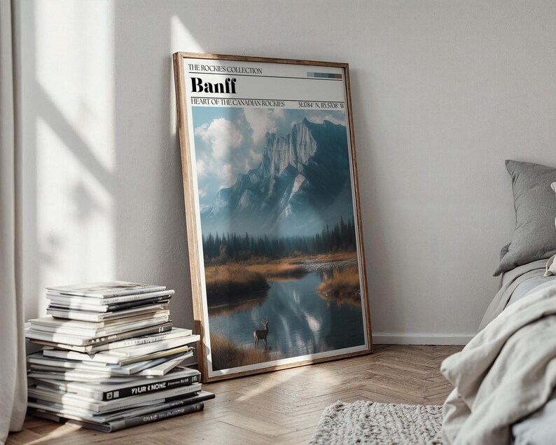 May include: A framed art print titled "Banff" from The Rockies Collection. The landscape art features a mountain range, a lake, and a deer. The print is in a wooden frame and leans against a white wall. A stack of magazines is in the foreground.