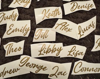 Könnte beinhalten: Eine Sammlung weißer Vinyl-Namensaufkleber mit goldfarbenen Buchstaben. Namen sind Emma, Ben, Ray, Judy, Keith, Denise, Emily, Alex, Lucas, Theo, Libby, Lisa, Drew, George, Zac und Connor. Die Aufkleber liegen auf einer dunkelgrauen Oberfläche.