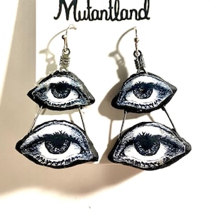 May include: A pair of silver dangle earrings with two black and white eye designs on each earring. The earrings are labeled "Mutantland".