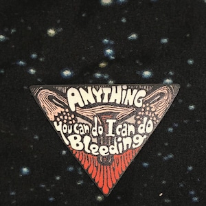 May include: A triangular patch with the words "ANYTHING You can do I can do Bleeding" in white and black. The patch has a red and white design at the bottom and a black background with white specks.