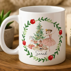 May include: White ceramic mug featuring a Christmas-themed illustration. The design includes a ballerina, Christmas tree, and the name "Sarah" in script. The mug is surrounded by a green wreath with red berries.