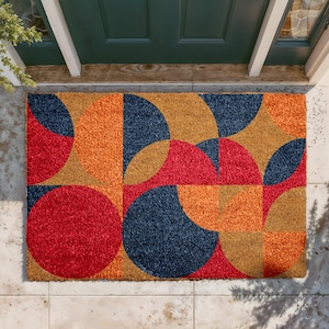 May include: Rectangular doormat with a geometric pattern of red, orange, navy blue, and tan shapes. The design features semi-circles and squares. The mat is made of a textured material and is placed in front of a green door.