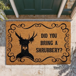 May include: A rectangular tan coir doormat featuring a black decorative scrollwork border. The mat displays a black silhouette of a knight with antlers and the text DID YOU BRING A SHRUBBERY? printed in a bold, black, serif font on the right side.