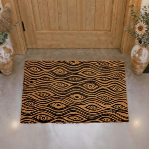 May include: A rectangular doormat with a repeating eye pattern in black on a tan background. The design features wavy lines that create the illusion of numerous eyes. The mat is placed in front of a wooden door, with decorative vases on either side.