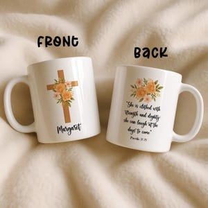 Könnte beinhalten: Zwei weiße Keramikbecher. Ein Becher zeigt ein Holzkreuz mit floralen Akzenten und dem Namen "Margaret". Der andere Becher zeigt ein Blumendesign und das Zitat: "Sie ist mit Stärke und Würde bekleidet; sie kann über die kommenden Tage lachen."