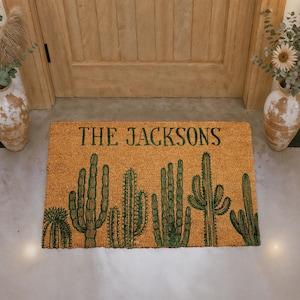 May include: A rectangular doormat with a natural coir background. The mat features a green cactus design and the text "THE JACKSONS" in a bold, dark green font. The design is centered on the mat.