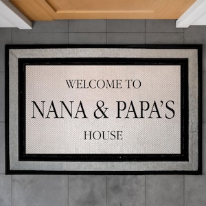 May include: Rectangular doormat with the text "WELCOME TO NANA & PAPA'S HOUSE". The mat has a white center with a textured pattern, framed by a black border and a gray outer edge. The mat is placed on gray tiles.