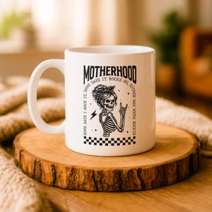 May include: White ceramic mug with the word "MOTHERHOOD" in black, along with a skeleton graphic and the text "SOME DAYS I ROCK IT, SOME DAYS IT ROCKS ME, EITHER WAY WE'RE ROCKIN". The mug is on a wooden coaster.