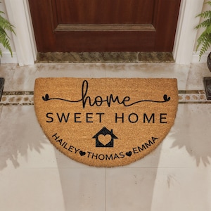 May include: A half-circle doormat made of natural coir fiber. The mat features the words "home sweet home" in black script, with a house and heart graphic. The names "Hailey", "Thomas", and "Emma" are also printed.