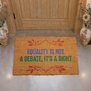 May include: A rectangular doormat made of natural coir fiber. The mat features the text "EQUALITY IS NOT A DEBATE, IT'S A RIGHT" in blue and green, with red decorative flourishes. The mat is placed in front of a wooden door.