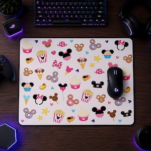May include: A rectangular mouse pad featuring a white background with a repeating pattern of colorful icons including Mickey Mouse ears, pink bows, popcorn buckets, ice cream cones, donuts, cupcakes, and yellow stars in various sizes and colors.