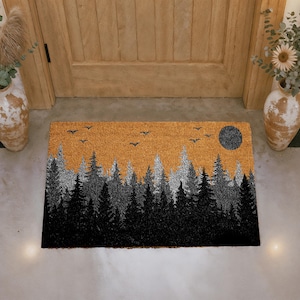 May include: A rectangular coir doormat featuring a forest scene. The design includes black and silver trees against an orange sky, with a dark gray moon and flying birds. The mat is placed in front of a wooden door.