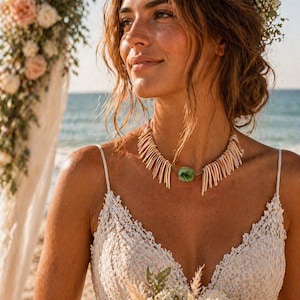 Op de afbeelding: Een vrouw draagt een witte kanten jurk en een unieke ketting met een groene stenen hanger. De ketting is gemaakt van beige en witte elementen. De achtergrond toont een strand en bloemdecoraties.