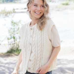以下が含まれることがあります： 木製ボタンが付いたクリーム色のケーブルニットベスト。ノースリーブセーターは、前面にテクスチャードケーブルデザインとリブ編みの裾が特徴です。ベストはボタンで留められ、明るい色の半袖シャツの上に着用されています。