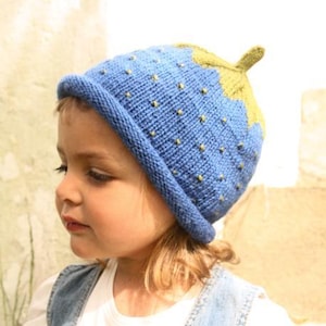 Könnte beinhalten: Eine blaue Strickmütze mit Erdbeermuster. Die Mütze hat eine blaue Basis mit gelben Punkten und eine grüne Oberseite und einen Stiel. Die Mütze wird von einem kleinen Kind getragen.