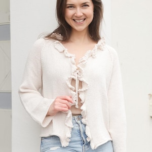 以下が含まれることがあります： フリルネックとフロントにリボンが付いた白いニットカーディガン。カーディガンは長袖でゆったりとしたフィット感です。