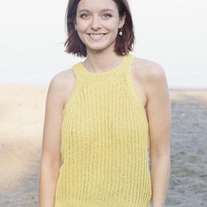 May include: A yellow knitted vest top with a high neckline. The top is made of a textured knit fabric and is paired with blue denim jeans. The image is taken outdoors with a blurred background.