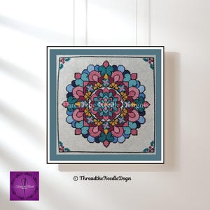 Pode incluir: Uma mandala bordada em ponto cruz, emoldurada, com um design complexo e simétrico. A obra apresenta um padrão circular central em tons de rosa, verde-azulado e dourado, rodeado por camadas de cores semelhantes. A moldura é verde-azulado suave e o fundo é bege claro.