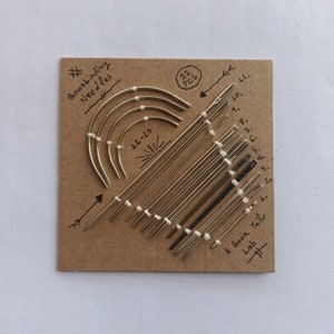 May include: A set of 22 bookbinding needles, arranged in a heart shape on a brown cardboard background. The needles are labeled with numbers 1 through 11 and 12-15. The text "Bookbinding Needles" and "A Book Tailor Lab" is written on the card.