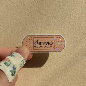 May include: A rectangular adhesive bandage sticker featuring a tan color with a holographic star pattern. The center displays the word brave enclosed in angle brackets with Morse code dots and dashes printed directly underneath the text.