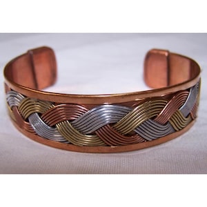 May include: A copper cuff bracelet with a woven design featuring bands of copper, silver, and gold-colored metal. The bracelet has a simple, open design and is displayed on a white surface.