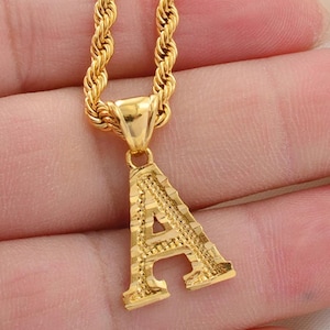 May include: A gold-colored necklace with a pendant in the shape of the letter "A". The pendant has a textured surface and is suspended from a gold rope chain. The necklace is displayed against a skin-toned background.