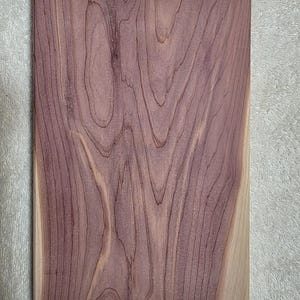 May include: A rectangular wooden board with a natural wood grain pattern. The board features a blend of purple and tan hues, with visible lines and swirls. The wood's texture is prominent, showcasing its natural characteristics.