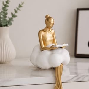 May include: A gold-colored figurine of a person with a hair bun sitting on a white, rounded cloud-like base. The figure is holding a small, open white book in their hands, with their legs dangling down from the cloud structure.