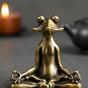 Può includere: Una statuetta di rana in bronzo in posizione yoga seduta. La rana ha grandi occhi rotondi ed è posizionata con le gambe incrociate e le braccia tese, le mani in un mudra. Un piccolo fiore di loto è alla base. Una candela accesa è sullo sfondo.