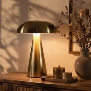 Peut inclure: Lampe de table dorée avec un abat-jour en forme de champignon et une base conique. La lampe est allumée, dégageant une lueur chaleureuse. Elle est posée sur un meuble en bois, avec un plateau, des bougies et un vase de fleurs séchées.