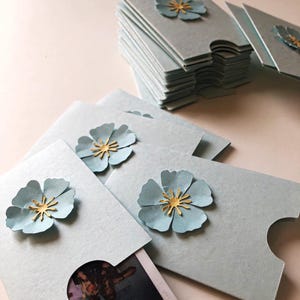 May include: Light blue paper photo holders with a die-cut edge and a light blue paper flower with a gold center. One holder contains a photo. A stack of similar holders is in the background.