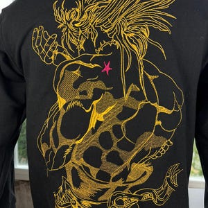 Pode incluir: Moletom preto com um grande desenho bordado em dourado nas costas. O desenho apresenta uma figura musculosa com cabelo espetado e uma estrela rosa. O bordado é detalhado e cobre grande parte das costas do moletom.