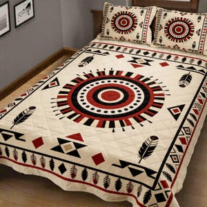 May include: Cream-colored quilt and pillow set with a geometric design in red, black, and tan. The quilt features a large circular pattern in the center, surrounded by geometric shapes and feather motifs. Two matching pillows complete the set.