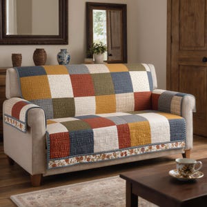 May include: A beige sofa with a colorful patchwork quilt cover. The quilt features squares of blue, yellow, red, green, and white. The sofa has rolled arms and a floral patterned trim. A small wooden table and a teacup are in front of the sofa.