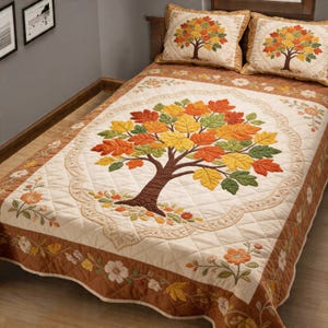 Könnte beinhalten: Eine gesteppte Tagesdecke und zwei Kissenbezüge mit einem gestickten Baum und Herbstblättern. Die Tagesdecke hat einen cremefarbenen Hintergrund, einen braunen Rand und florale Akzente. Die Kissen haben das gleiche Baumdesign.