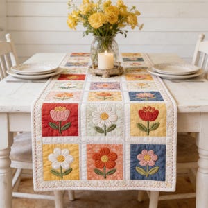 May include: A floral quilted table runner with embroidered flower blocks in various colors, including red, yellow, and blue. The runner is placed on a white table, with a vase of yellow flowers and a lit candle in the background. The table is set with plates.