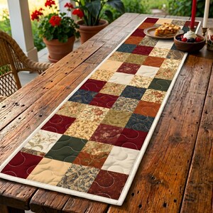 May include: A patchwork table runner with a variety of patterns and colours, including burgundy, beige, and green. The runner is placed on a rustic wooden table. The runner's design adds a touch of country charm to the setting.